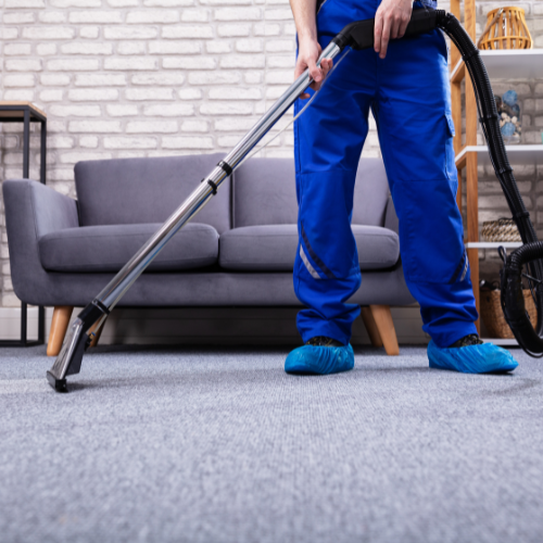 Person in blue overalls vacuuming a gray carpet in a living room.
