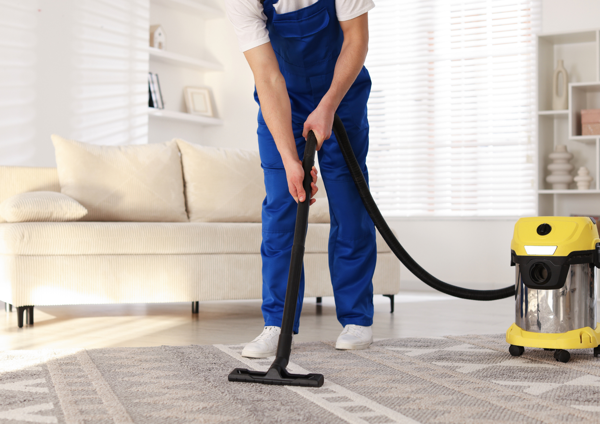 Person in blue overalls vacuuming a patterned rug in a bright room with a couch and shelving.