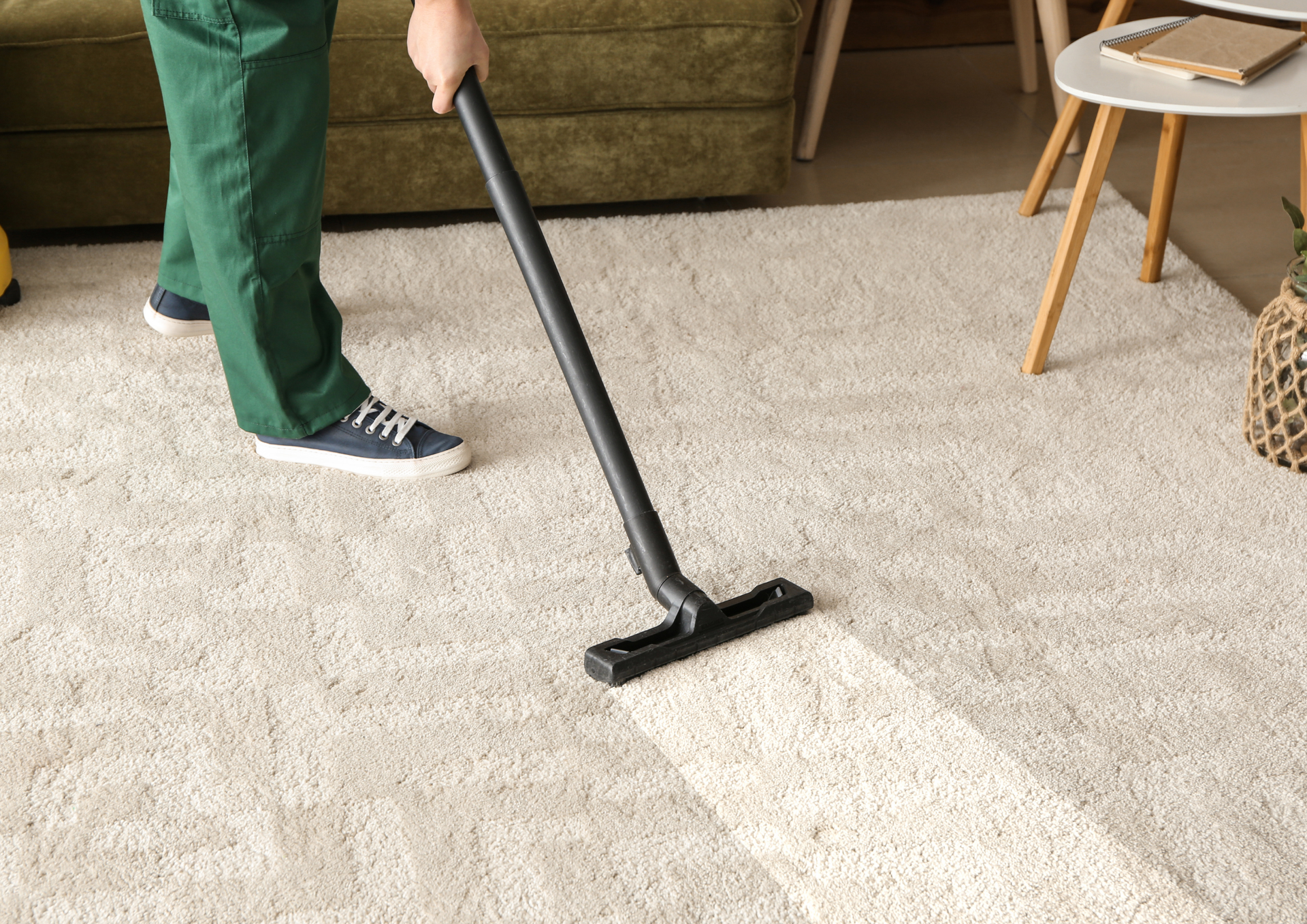 Person vacuuming a cream-colored rug, leaving a clean trail. In a living room setting, near a sofa and side table.