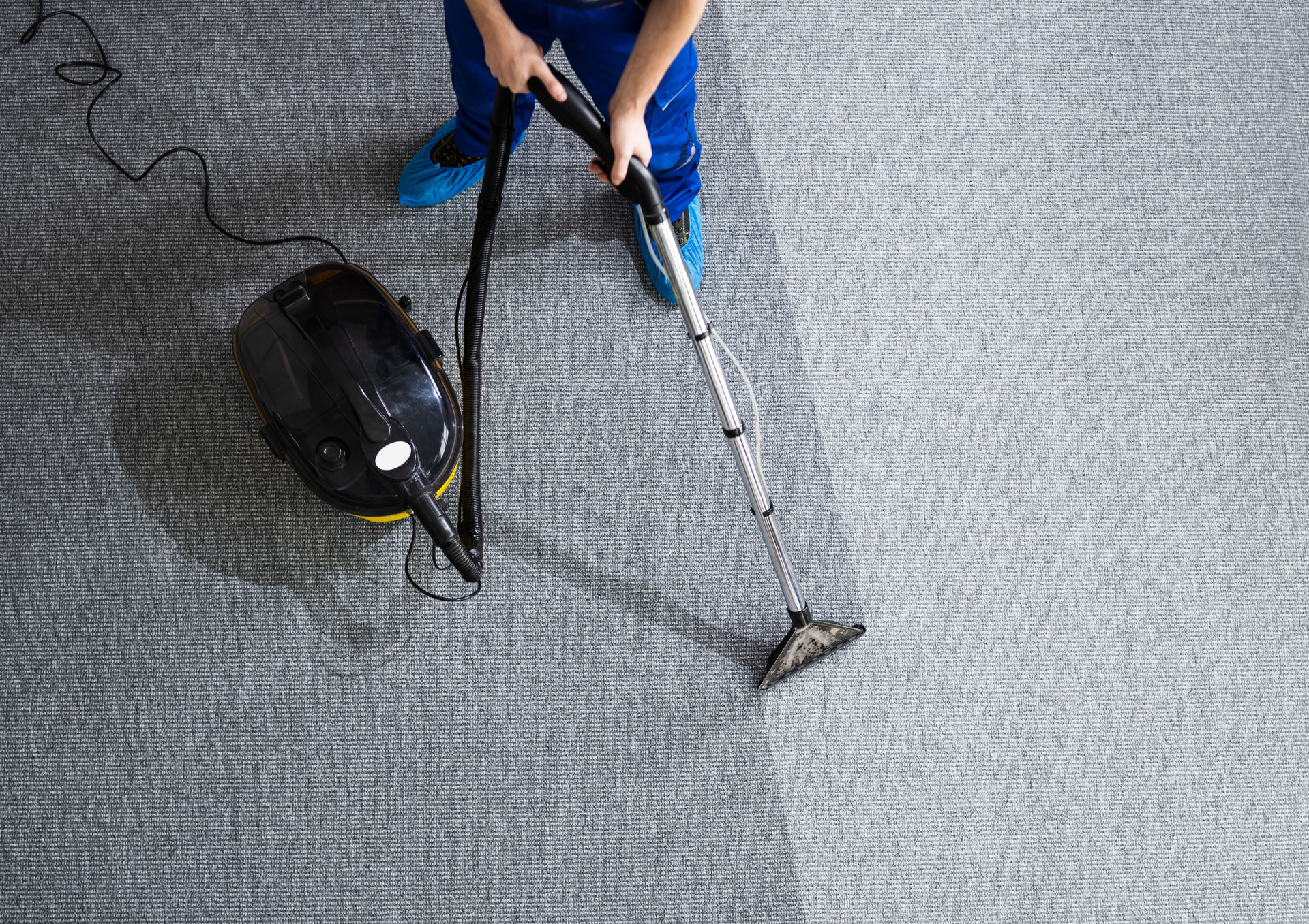 Person in blue overalls steam cleaning a gray carpet with a black machine.