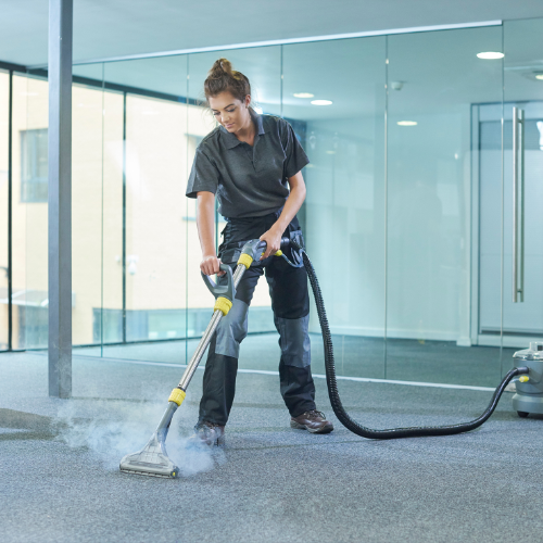 Woman steam-cleaning carpet in an office. She's holding a machine, and steam rises from the cleaner.