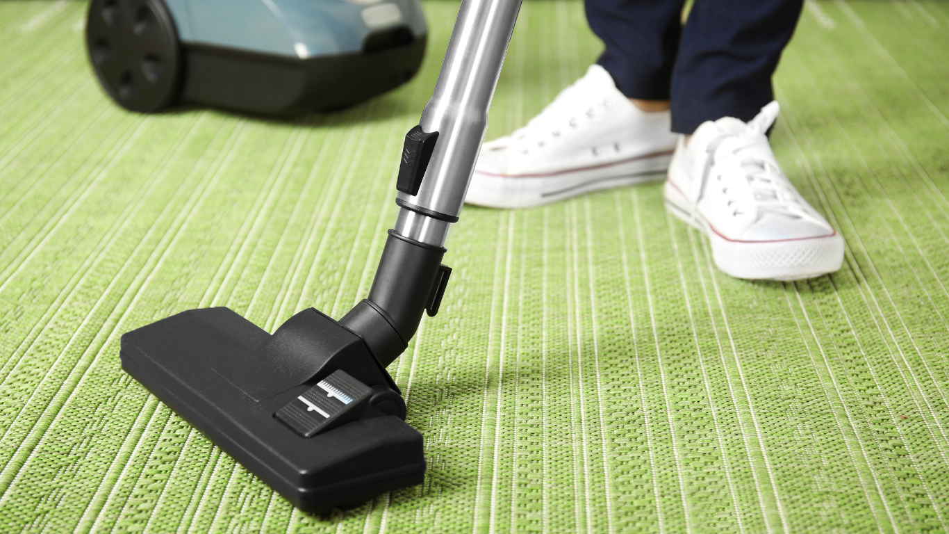 Vacuum cleaner cleaning a green carpet. Person in white sneakers stands nearby.