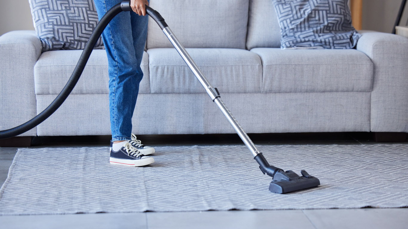 Person vacuums a rug in front of a gray couch, wearing jeans and sneakers.