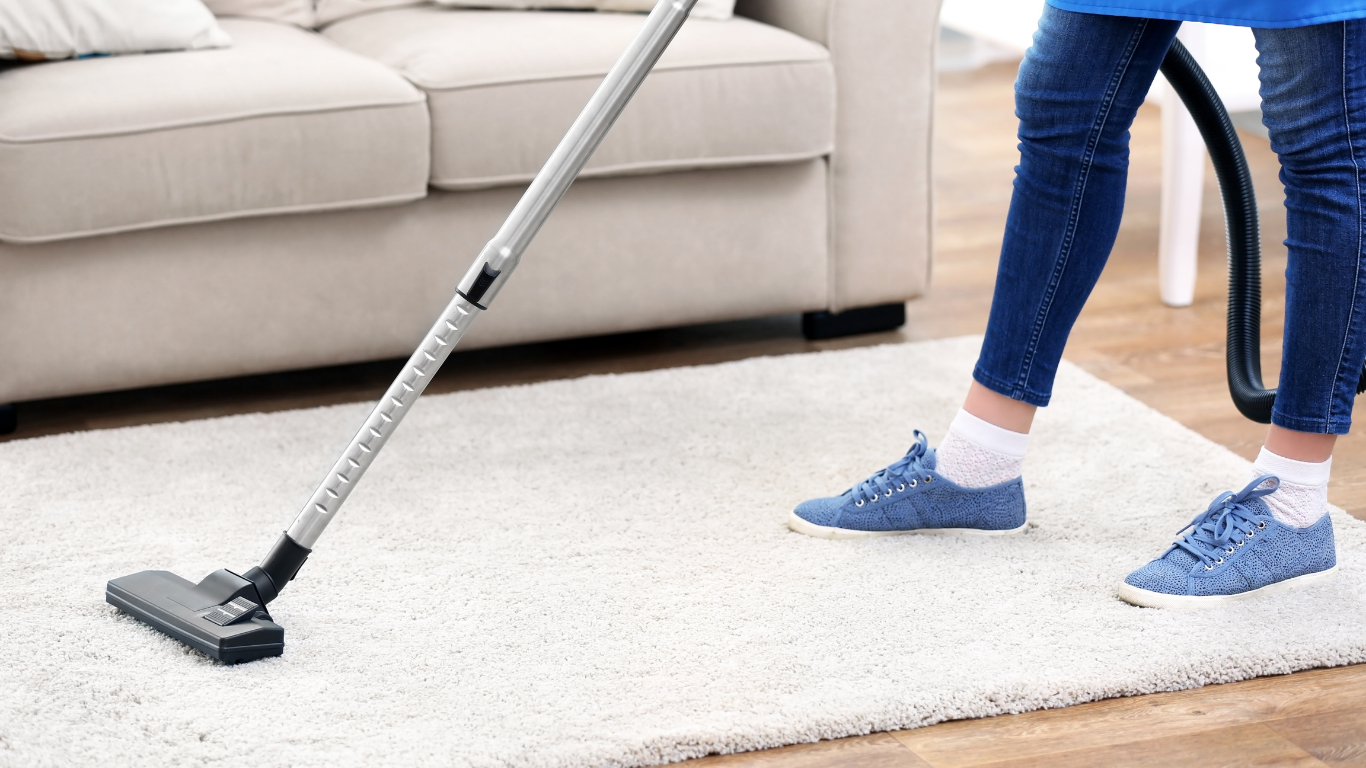 Vacuum cleaner cleaning a white fluffy rug near a beige couch. A person in blue jeans and shoes vacuums.