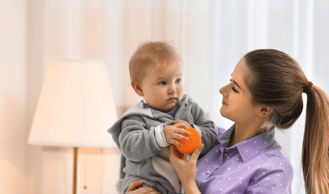 Woman holding baby indoors, baby looking away, holding orange toy. Lamp in background.