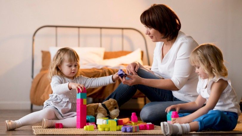 Woman and two young children playing with colorful building blocks on a rug.