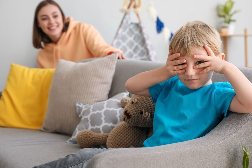 Child with hands over eyes, playing peek-a-boo, while a woman smiles behind him on a couch.