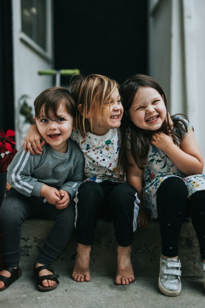 Three smiling children with arms around each other, sitting on a step.