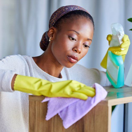 Person cleaning a wooden cabinet with yellow gloves and a purple cloth, holding a spray bottle