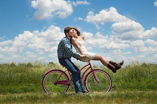 A couple kissing while sitting on a pink bicycle in a grassy field.