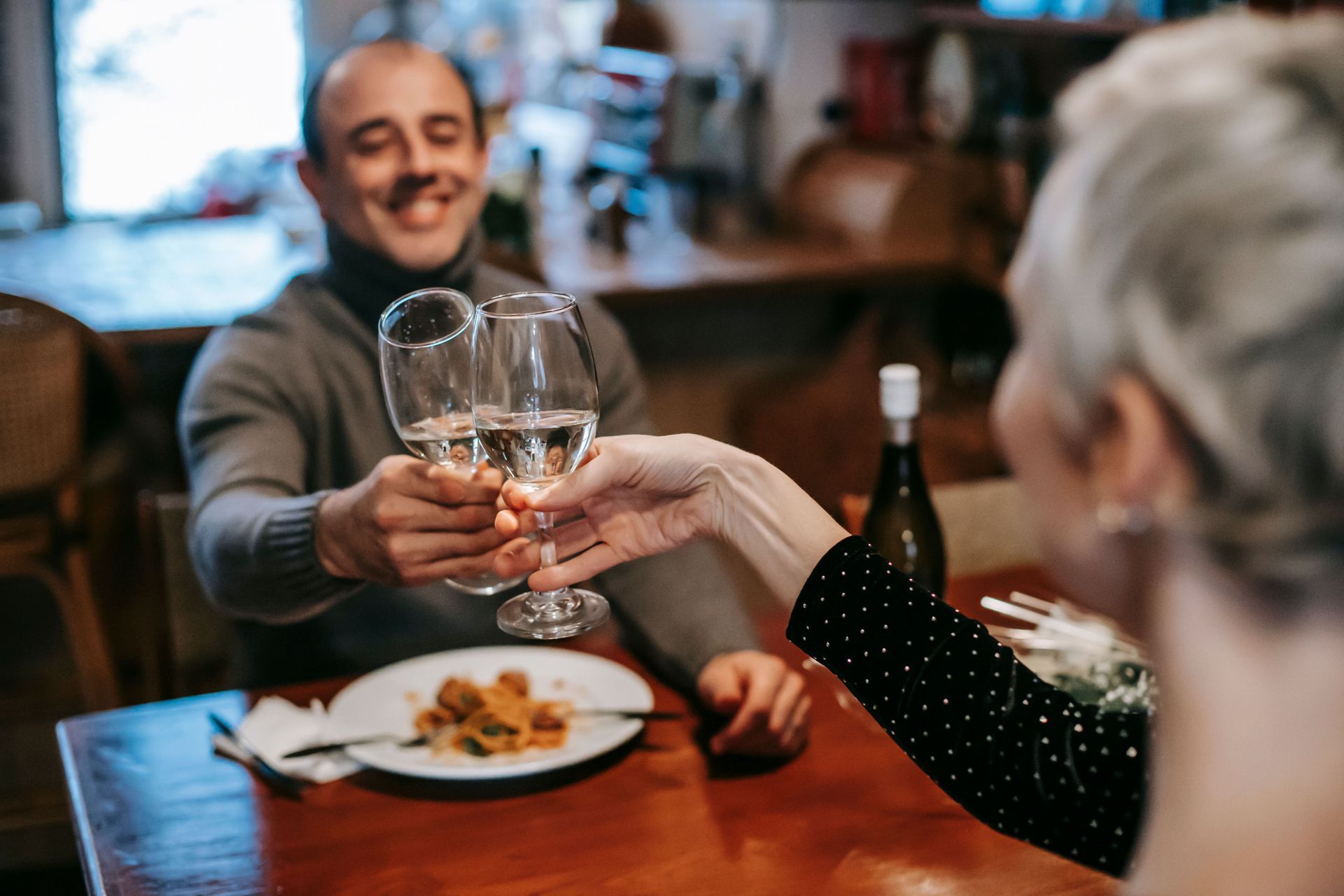 Two people clink wine glasses at a dining table with a plate of food and a bottle in a cozy setting.