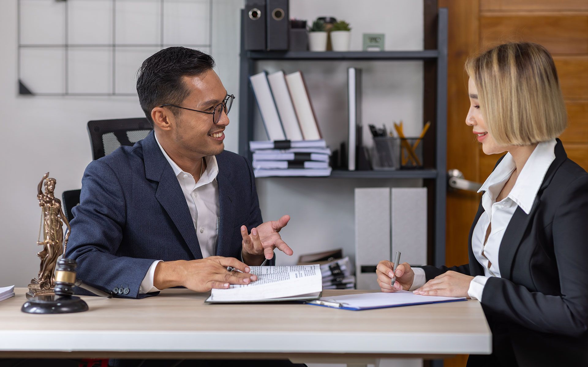 Man in suit gestures, talking to woman in suit as she writes at a desk. Office setting.