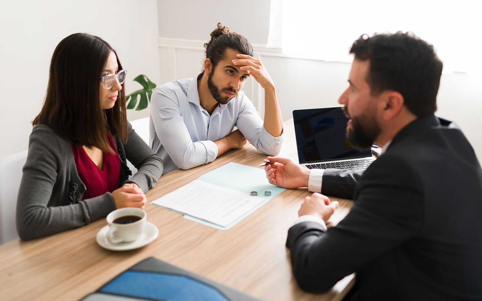 Couple looking stressed, meeting with a suited professional at a table with documents, coffee, and laptop.
