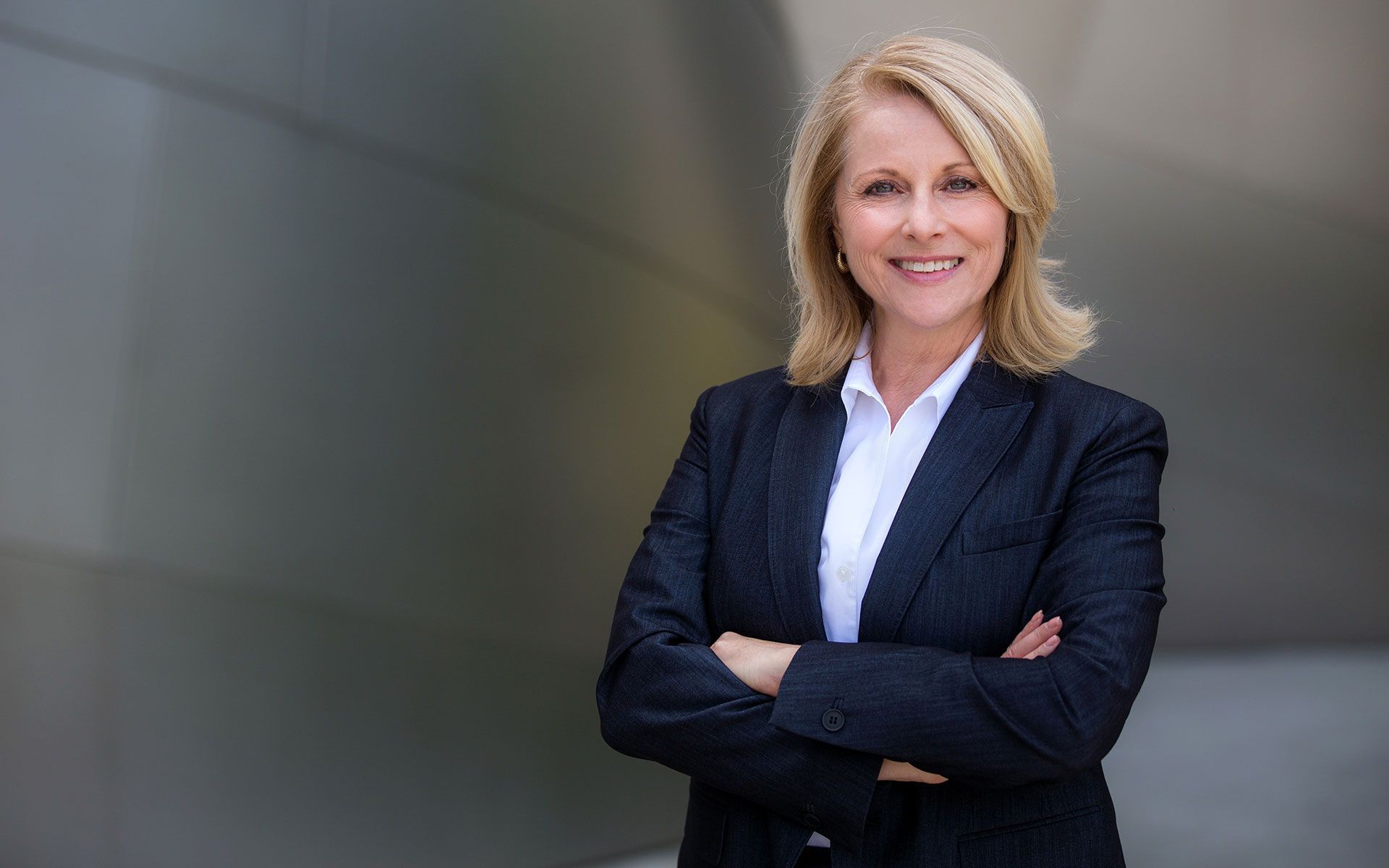 Blonde woman in business suit with arms crossed, smiles at camera. Set against a modern, metallic building.