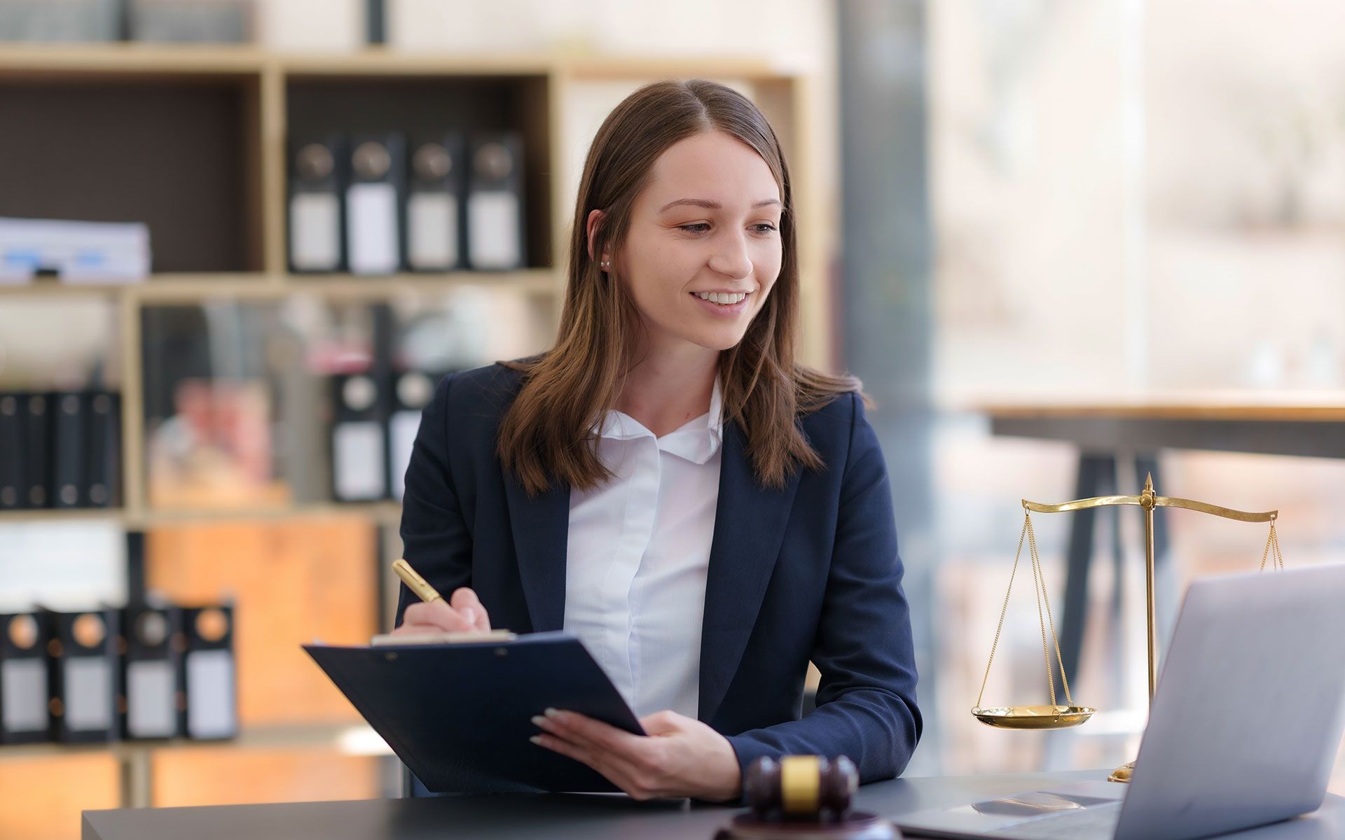 Woman lawyer smiles, takes notes, and works at desk with laptop, gavel, and scales of justice.