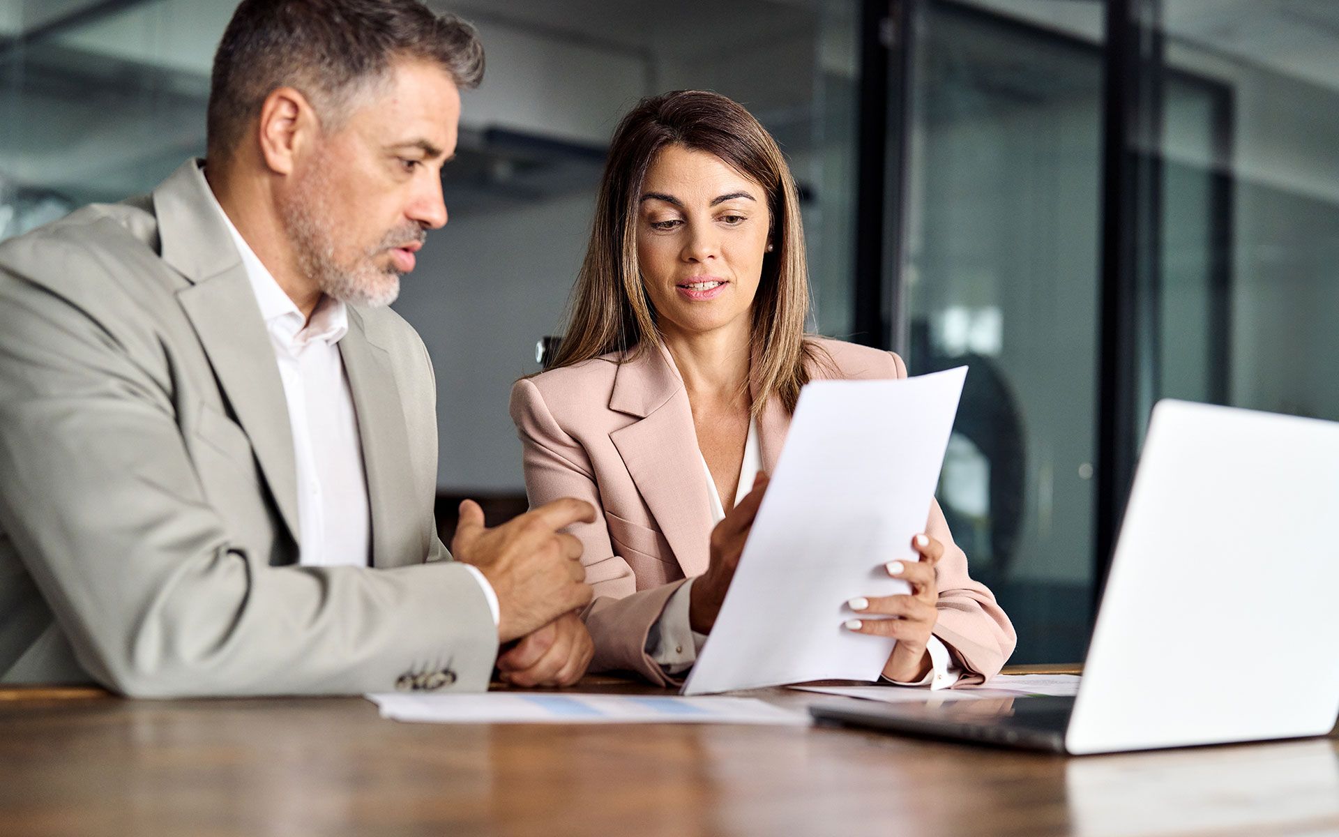 Man and woman in business attire review documents at a table with a laptop.