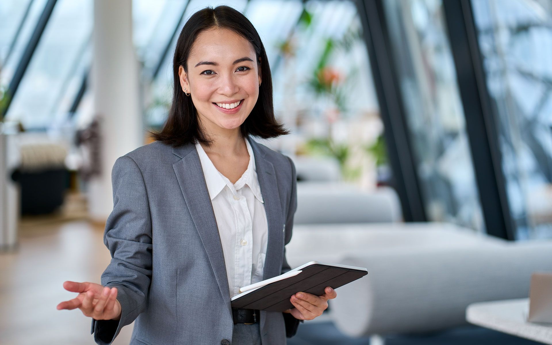 Asian woman in a gray suit, smiling and holding a tablet in an office setting.