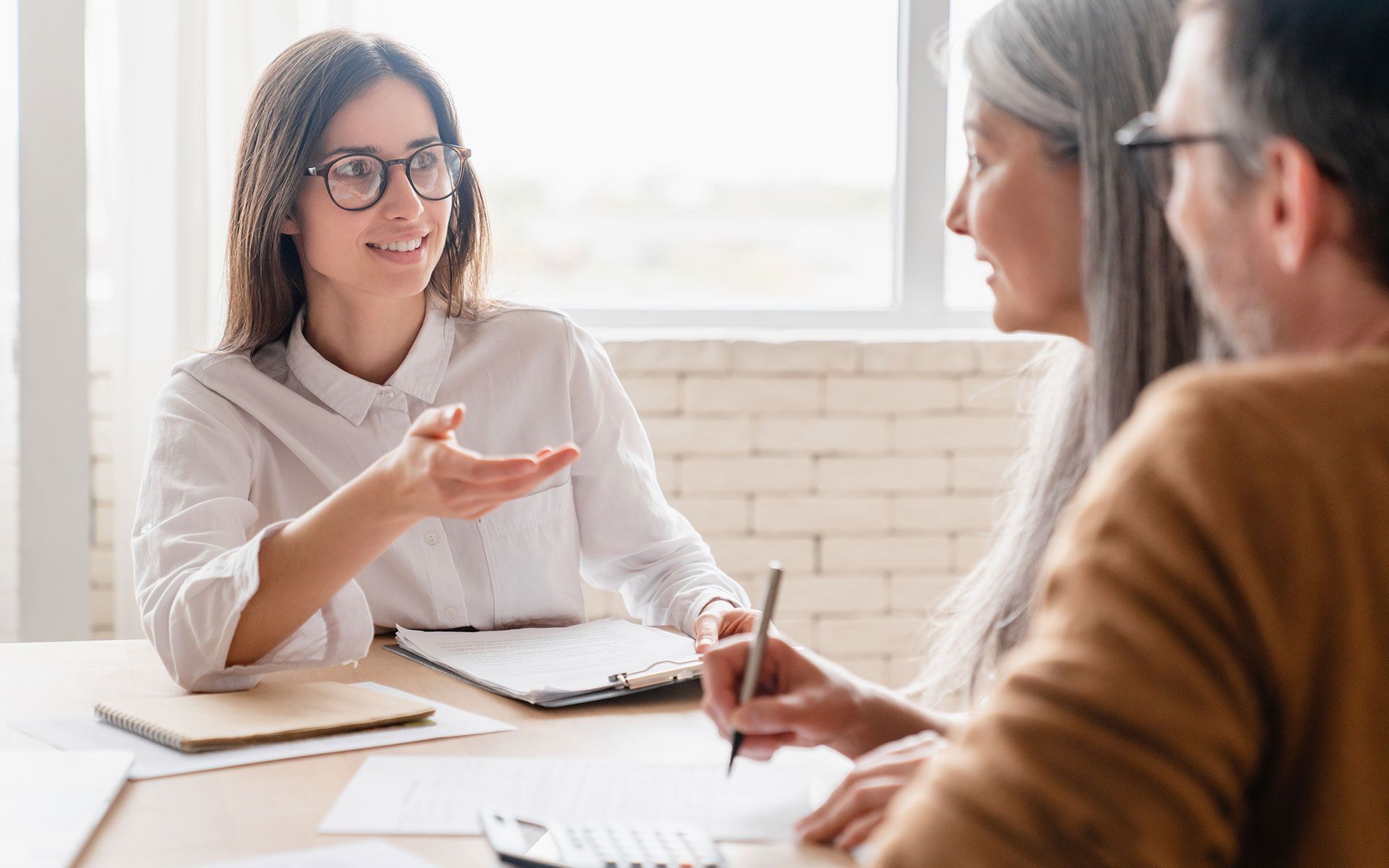 Woman in glasses gestures while discussing paperwork with older couple at a table.