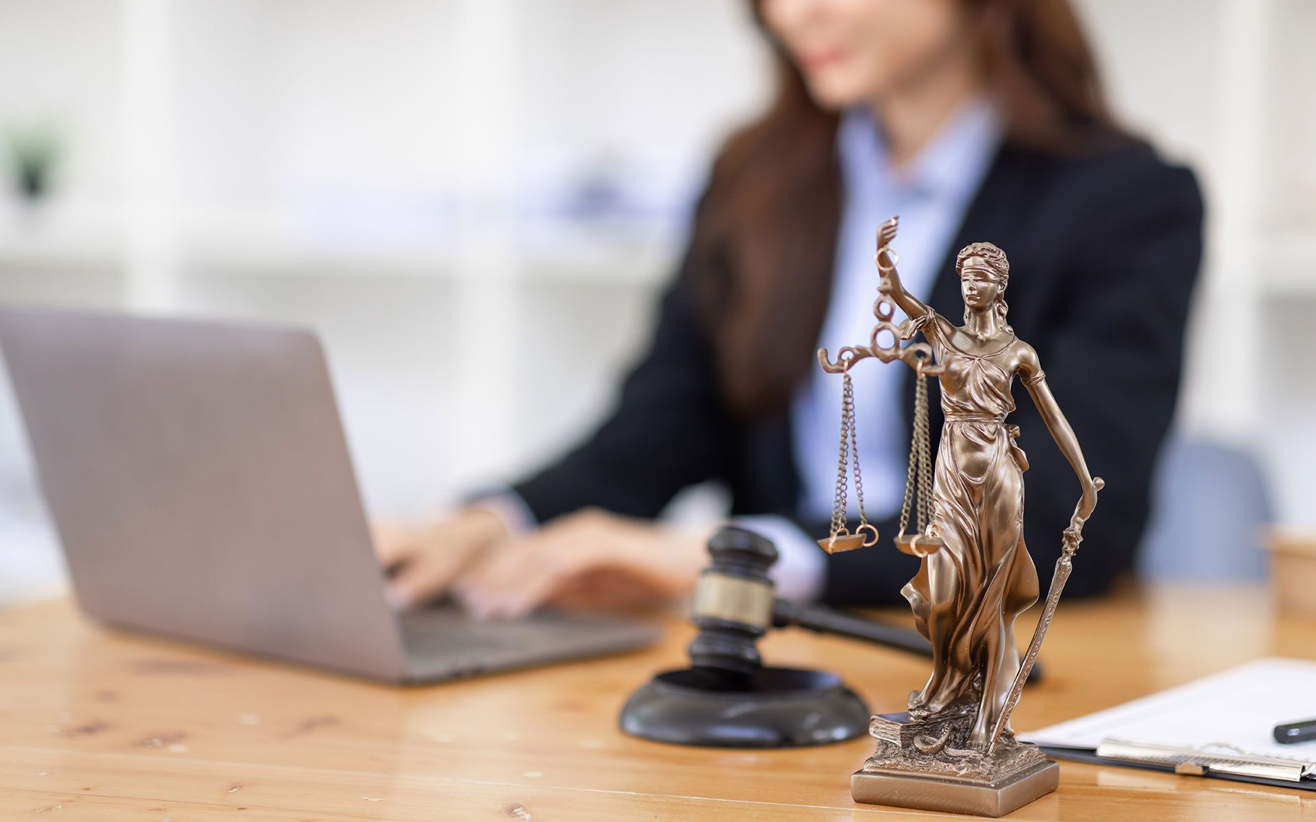 Woman in suit working on laptop with a statue of Lady Justice and gavel on a wooden desk.