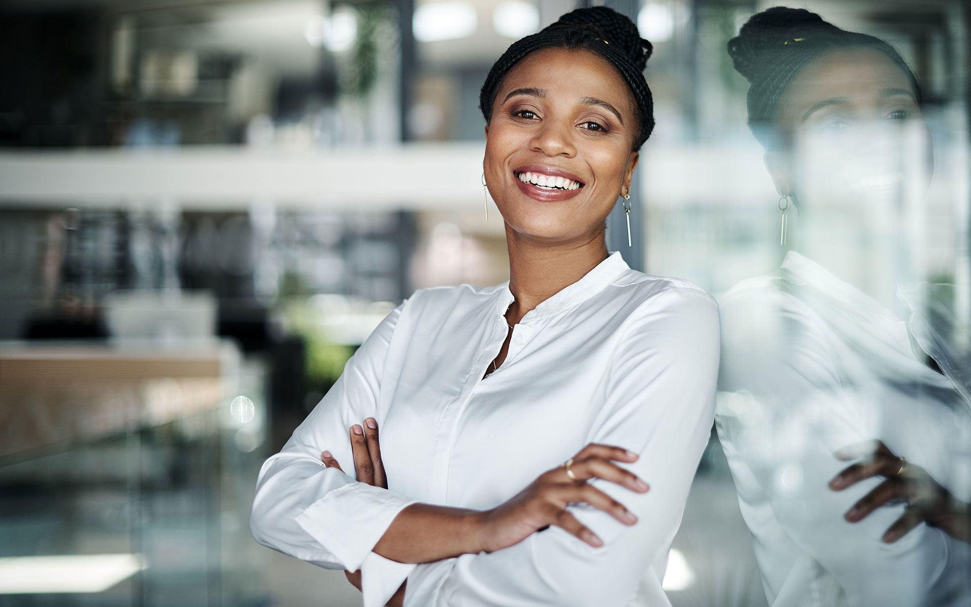Woman in white shirt smiling, arms crossed, leans against glass wall in an office.