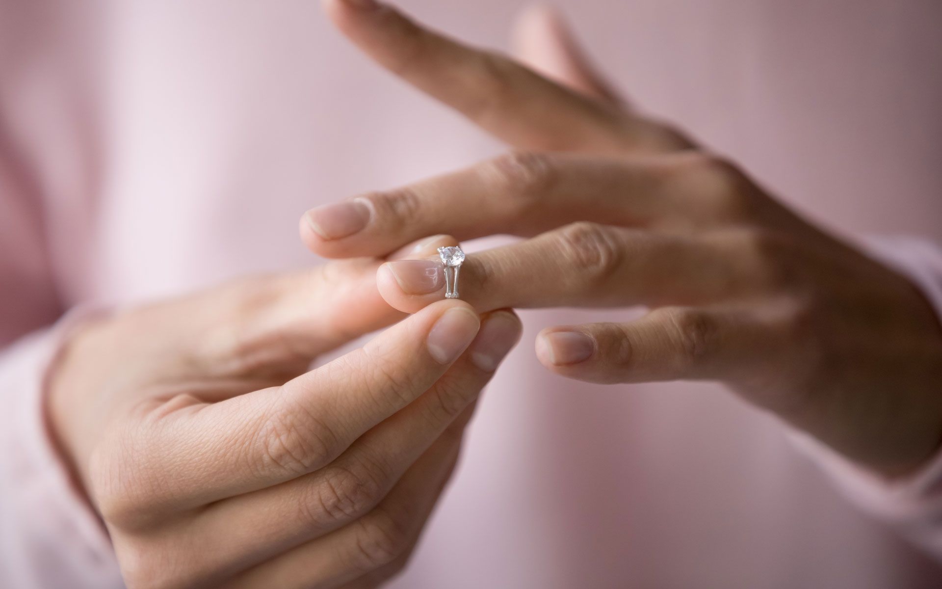 Person's hands removing a diamond ring from their finger. Close-up shot, light pink shirt.