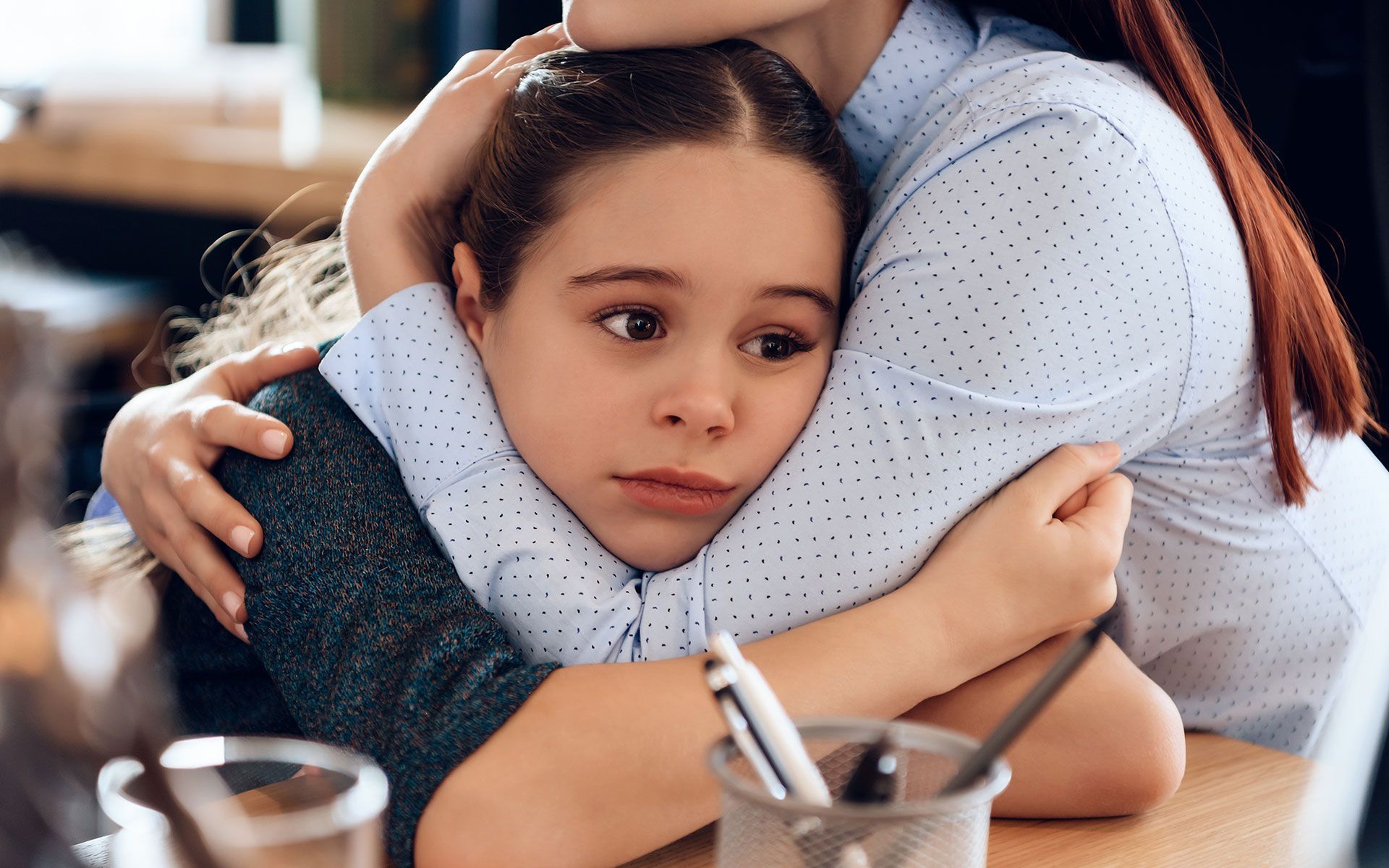 A woman comforts a worried girl, hugging her at a table; they are in an office setting.