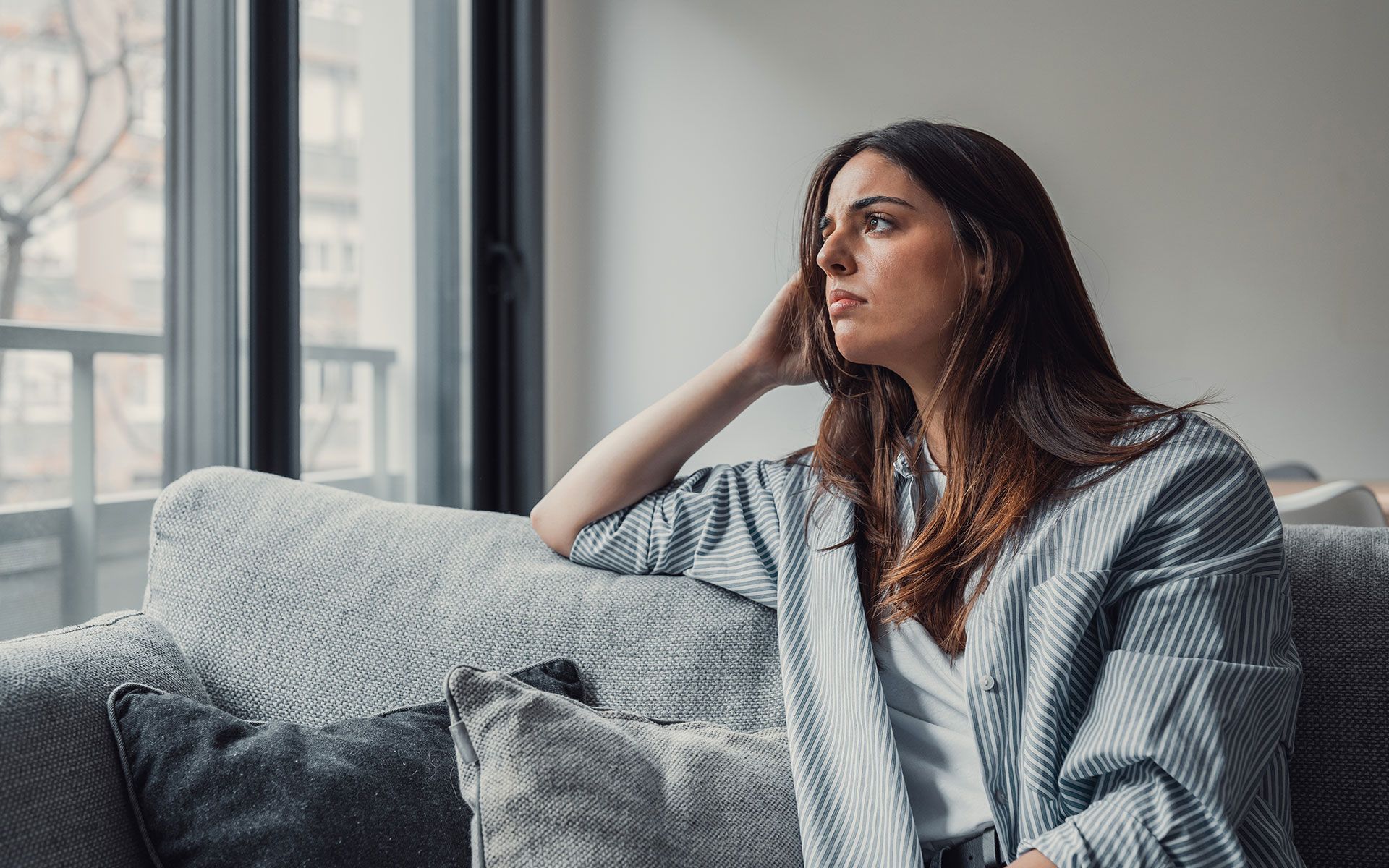 Woman with brown hair, looking out window, resting on a gray sofa, wearing a blue striped shirt.