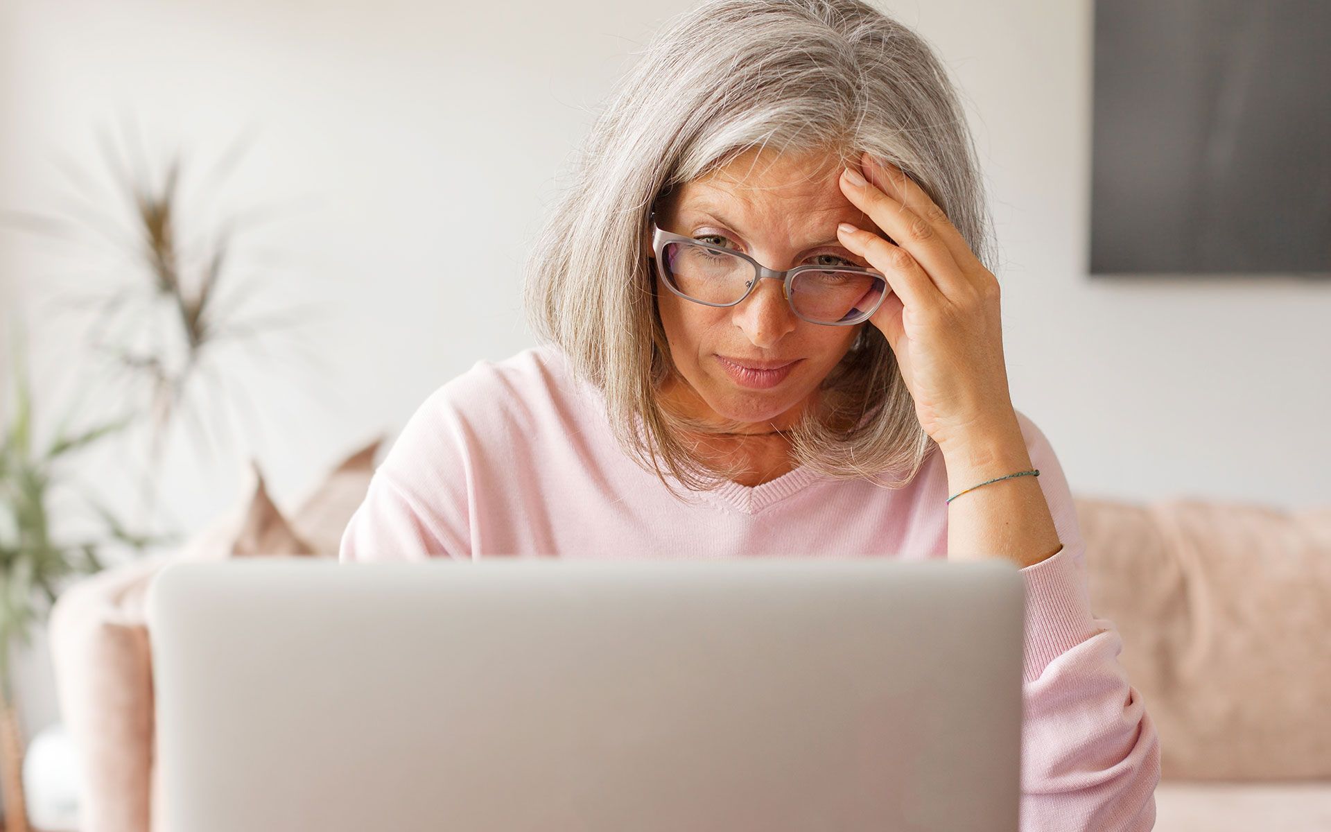 Woman with grey hair, glasses, looking intently at a laptop screen, hand on forehead, concerned expression.