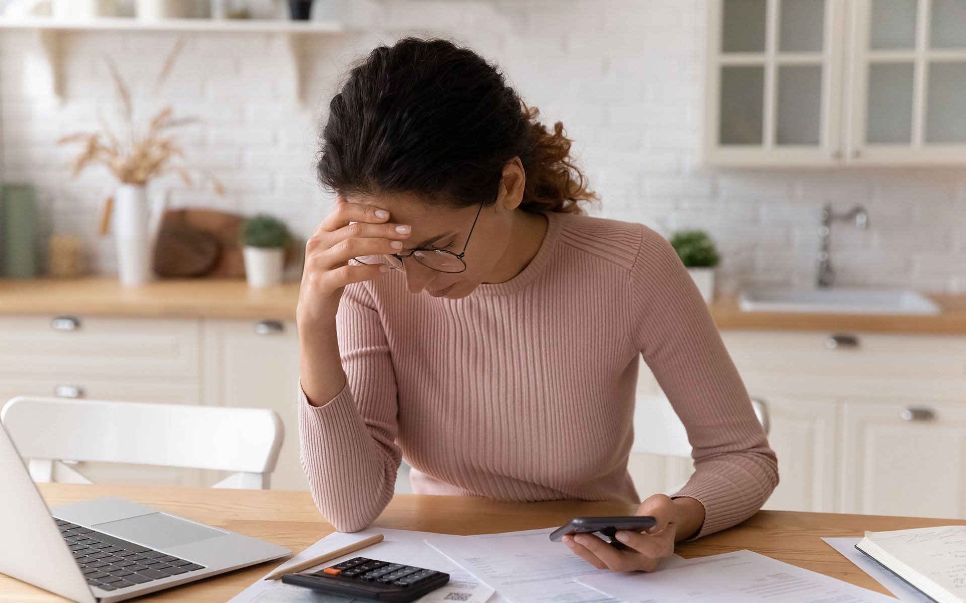 Woman in a kitchen, stressed, holding head, looking at bills with a calculator and laptop.