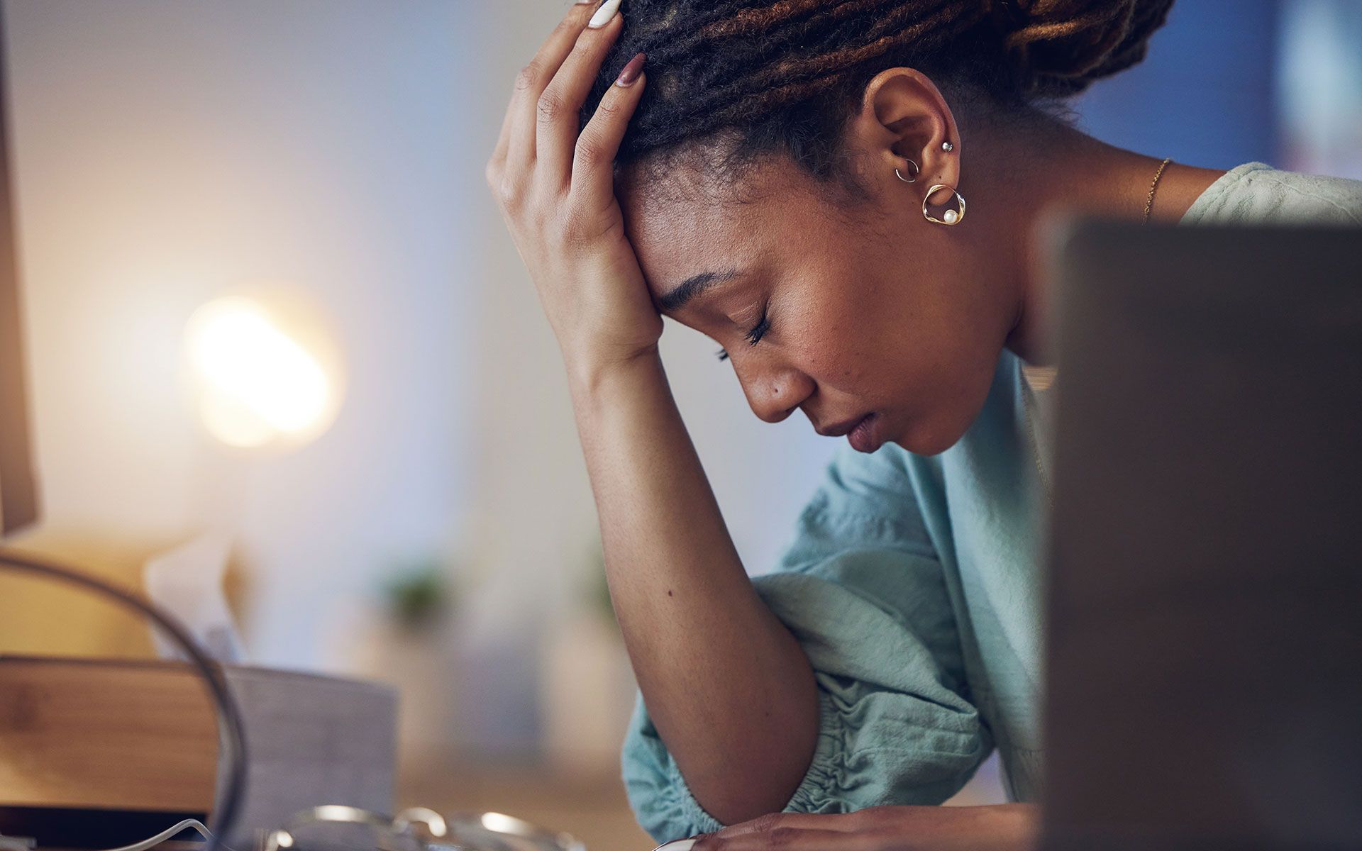 Woman with head in hand, stressed, at desk with laptop and lamp.