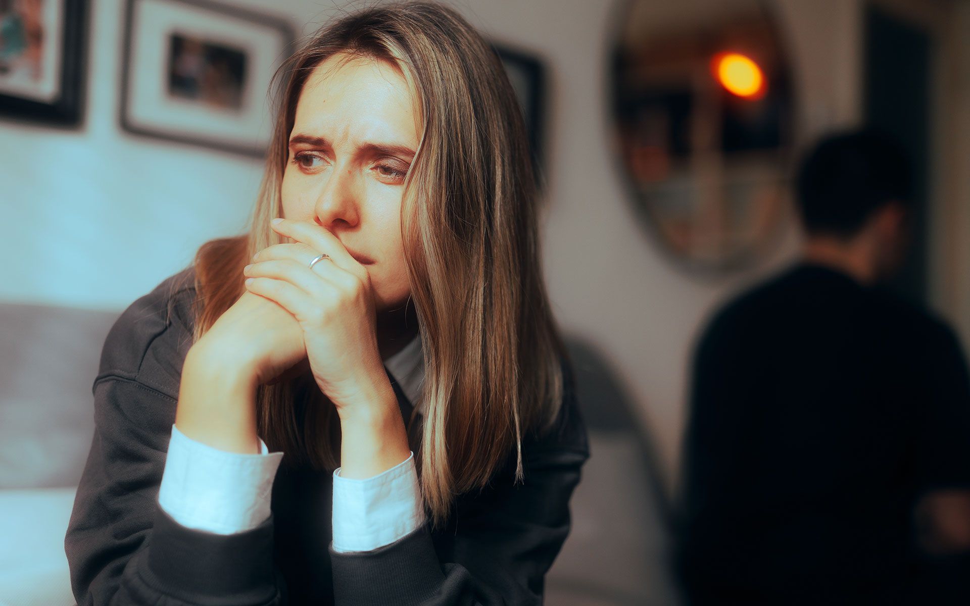 Woman with worried expression, hands clasped to her face; man in background, blurred, turned away.