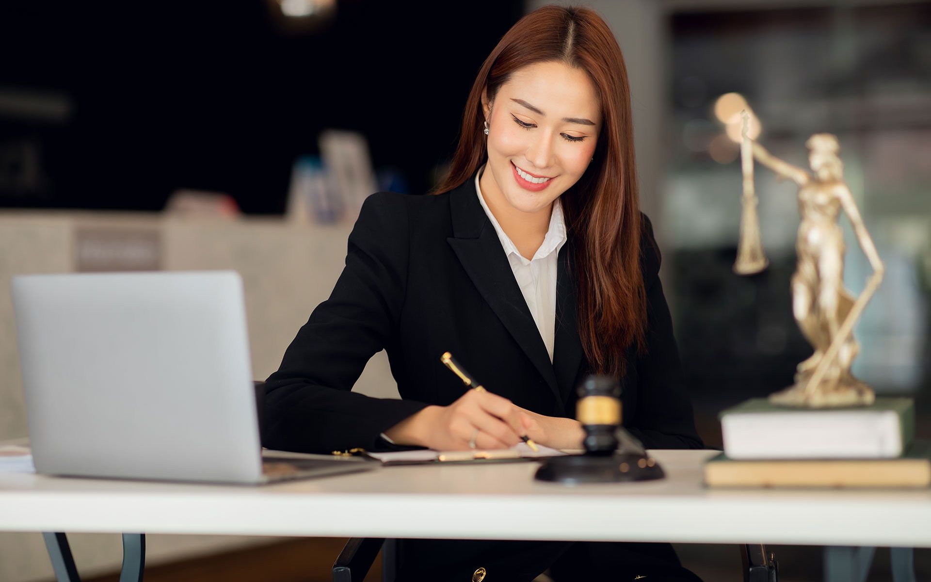 Asian woman lawyer in a black suit, smiling and writing at her desk with a laptop and law books.