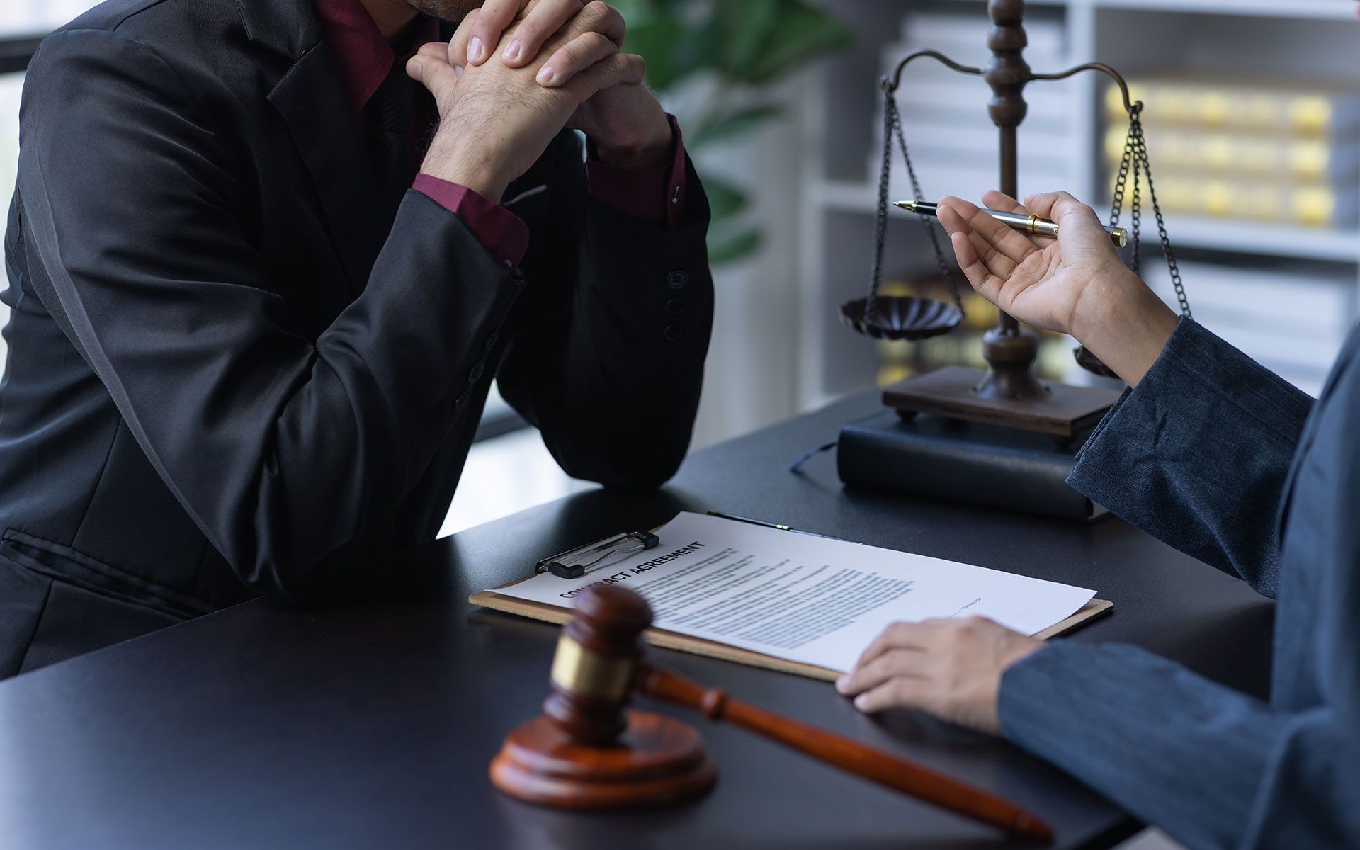 Person in suit consults with lawyer, reviewing document. Gavel and scales of justice on desk.