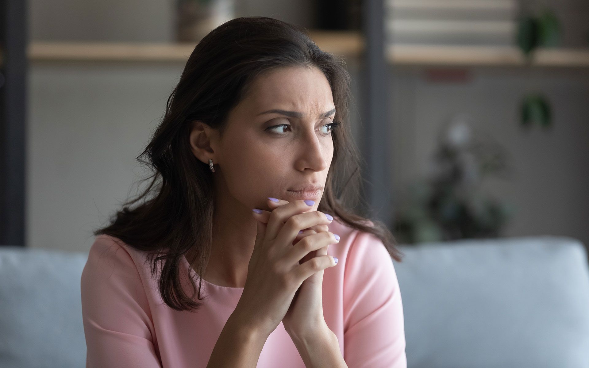 Woman with hands clasped, looking thoughtful, wearing a pink shirt, indoors.