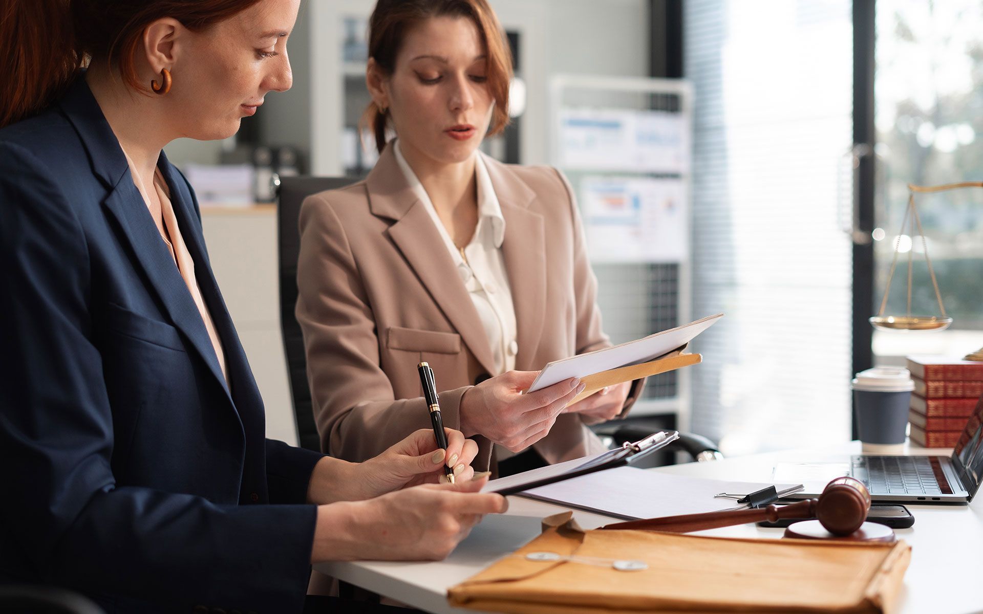 Two women in a law office review documents. One writes, the other holds papers.