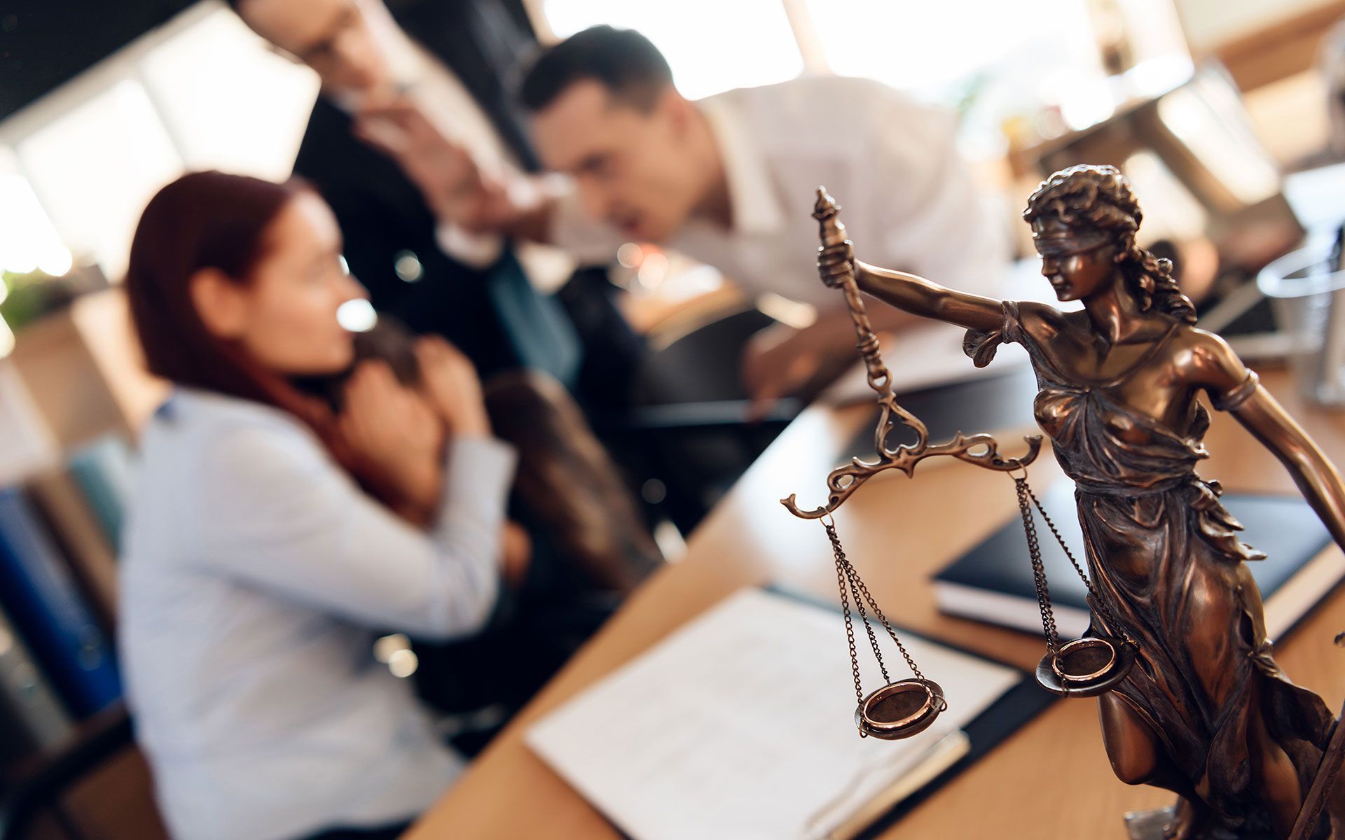 Statue of Lady Justice on desk; lawyers in discussion at a table in an office.