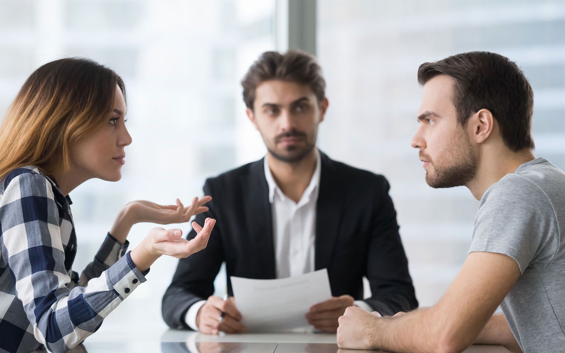 Woman arguing with man in office setting, mediator holds papers.