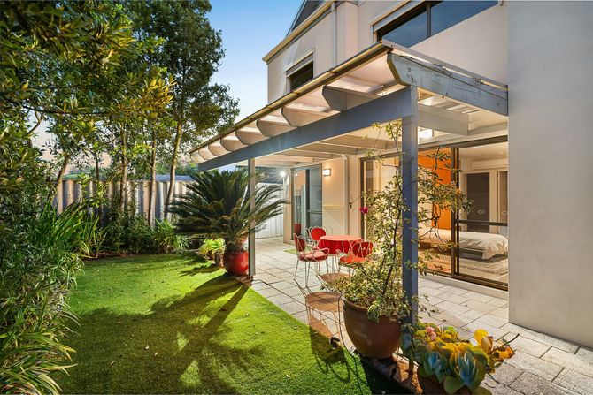 Sunlit backyard patio with glass awning, red chairs, potted plants, and a green lawn beside a white house