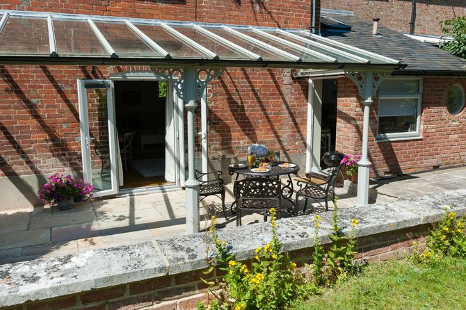 Brick patio with glass-roofed pergola, open doors, and outdoor table and chairs by a garden border