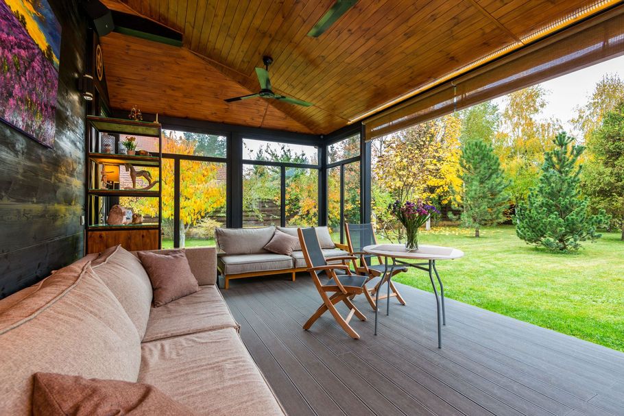 Covered patio with beige sofas, wooden chairs, and a round table opening to a green garden.