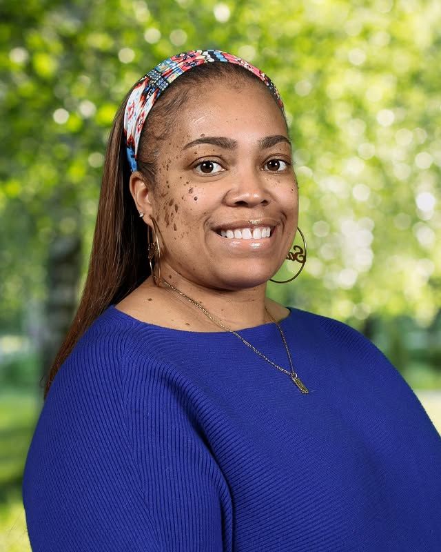 Smiling person with long hair and a colorful headband, wearing a blue sweater and gold jewelry against a green background.