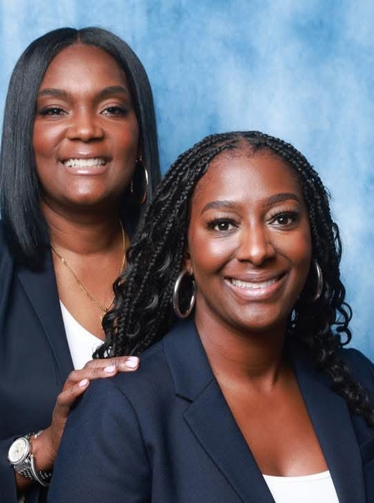 Two women in professional navy blazers smile against a blue textured backdrop, one resting a hand on the other's shoulder.
