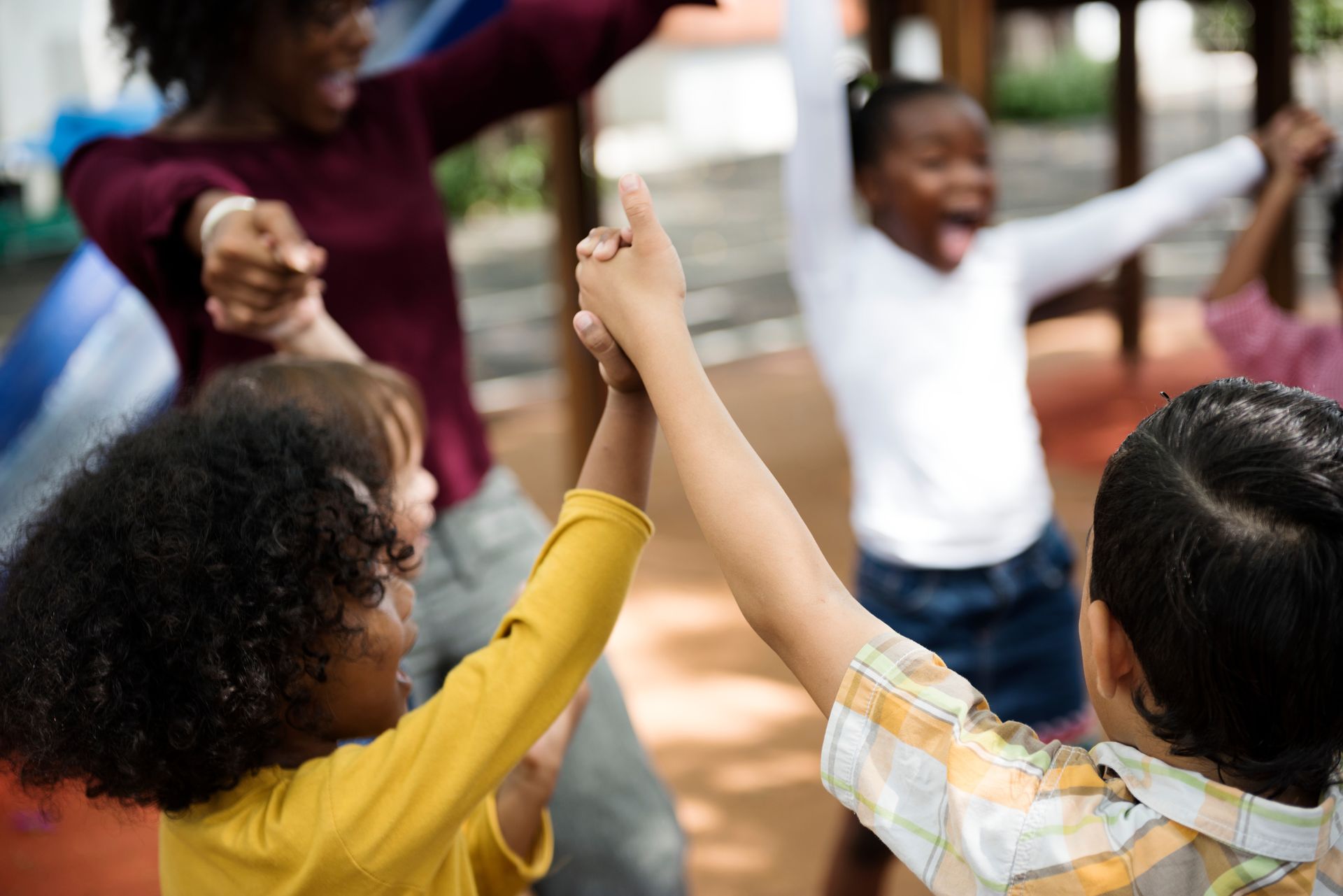 A teacher and a group of children laughing and holding hands in a circle on a playground.