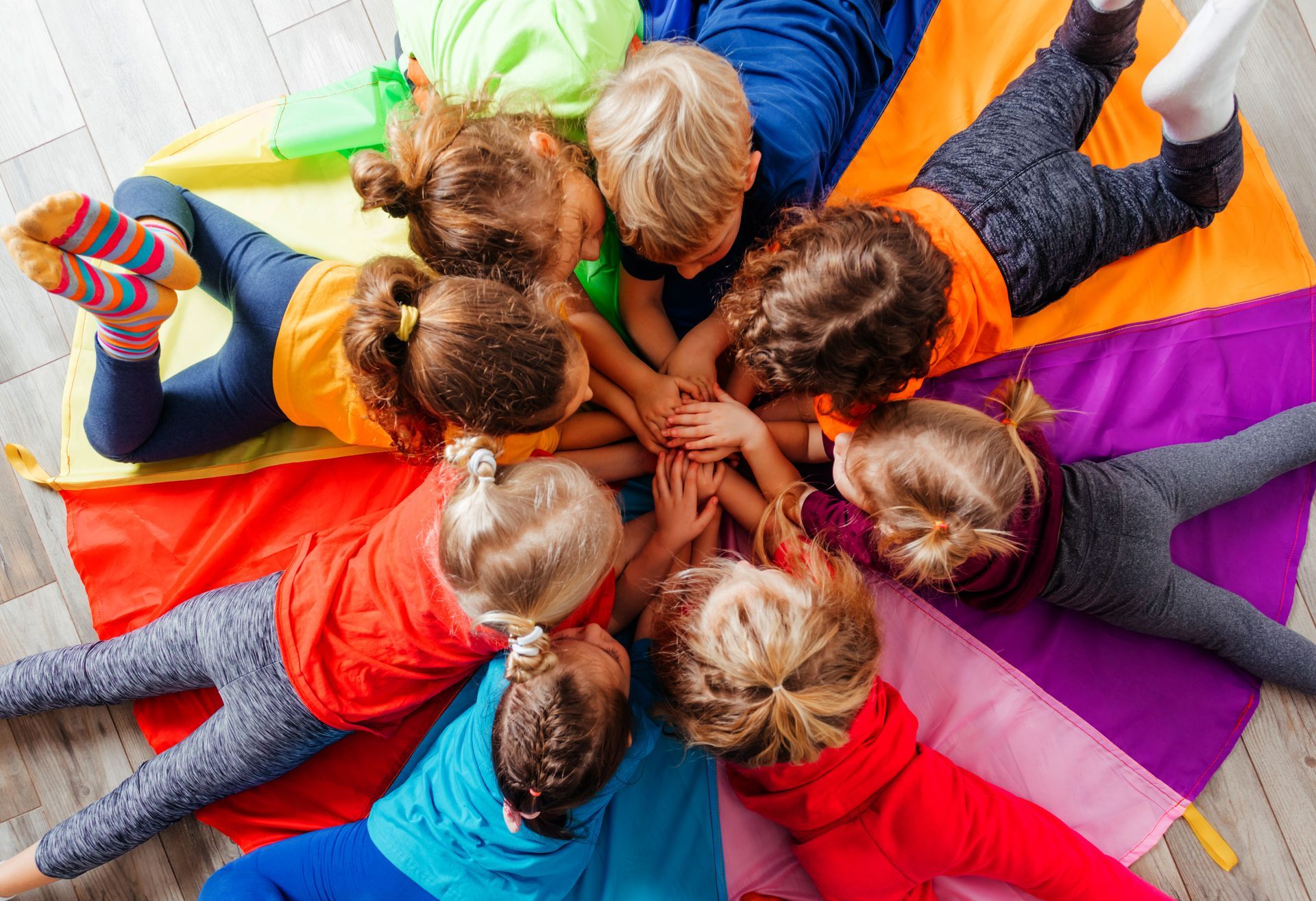 A group of children lying in a circle on a multicolored mat, all reaching toward the center to stack their hands together.