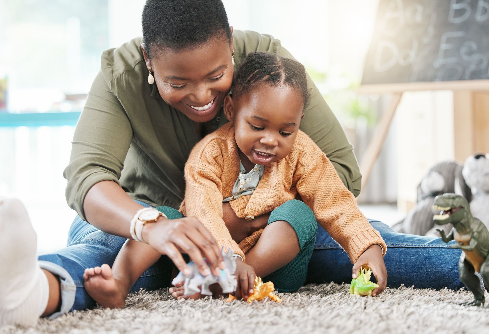 An adult and child sit on a carpet, smiling and playing with plastic toy dinosaurs in a room with a chalkboard.