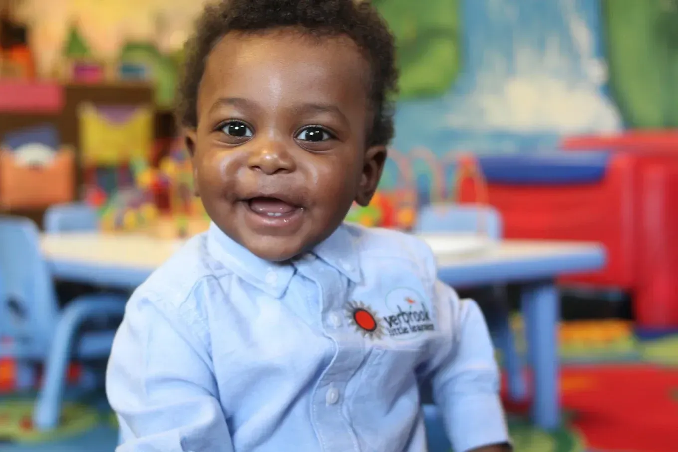A smiling baby wearing a light blue button-down shirt with a logo in a bright, colorful classroom.