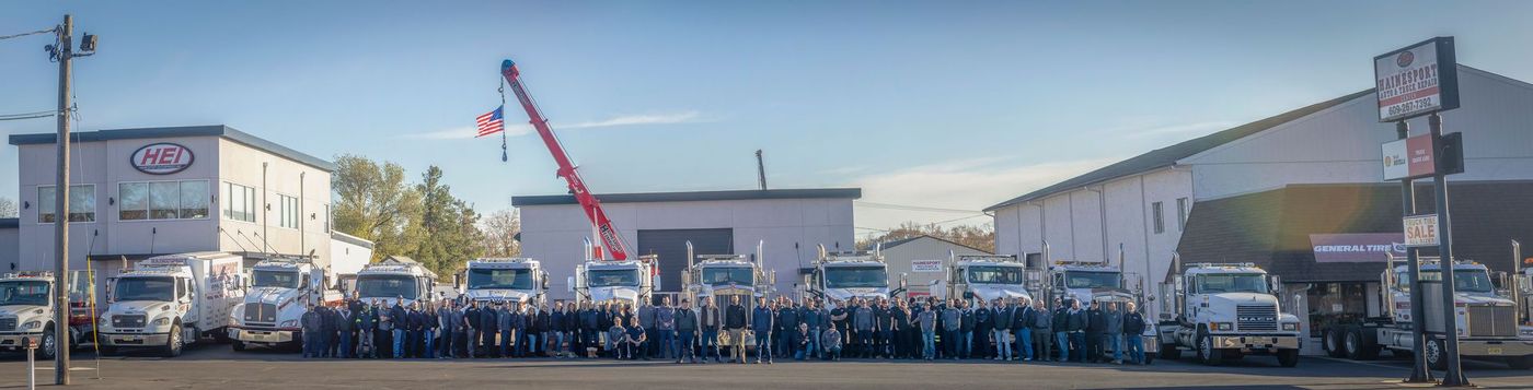 Group of people standing with trucks in front of buildings on a sunny day. A crane and sign are visible.