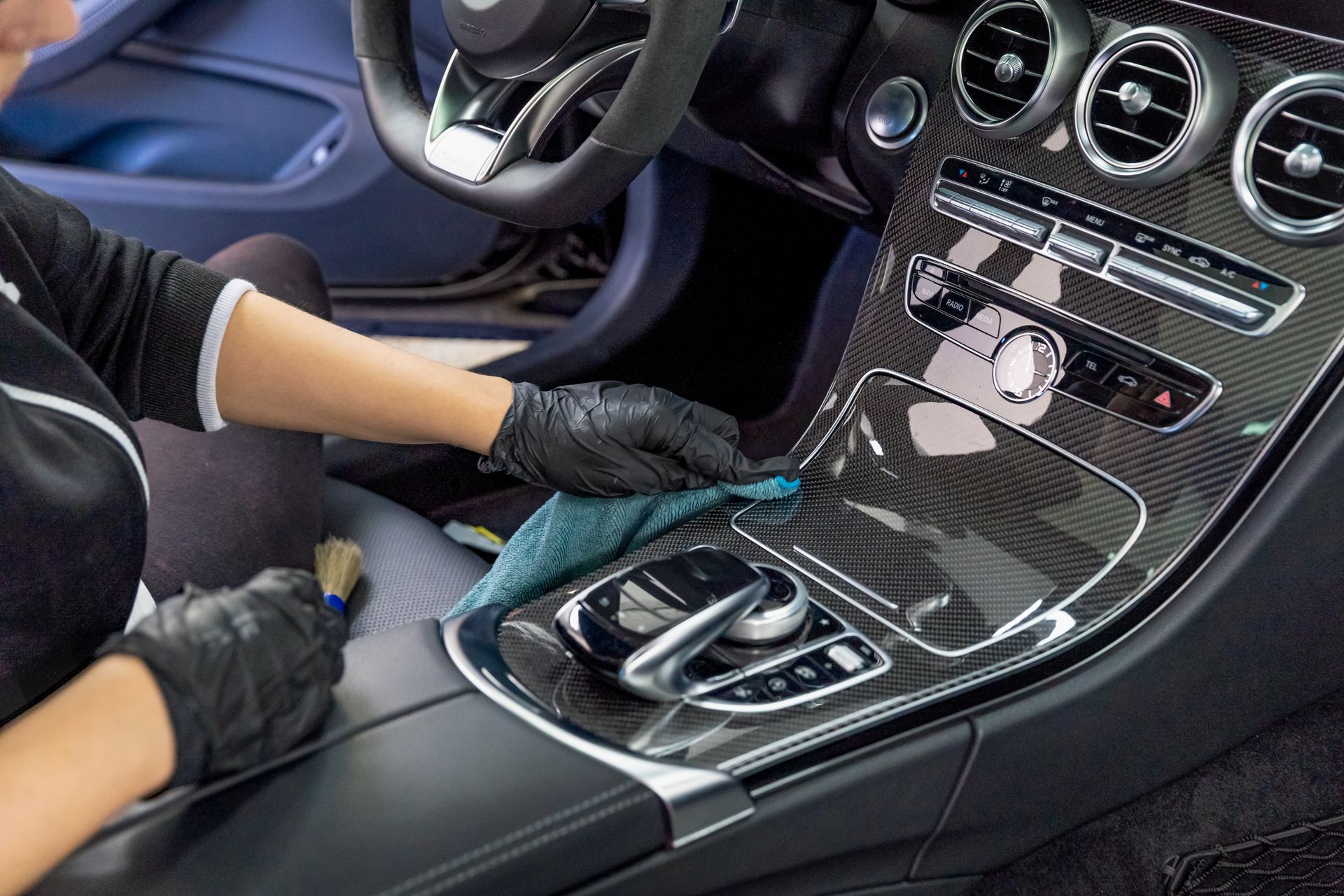 Person wearing black gloves cleaning a car's dashboard with a microfiber cloth and a brush.
