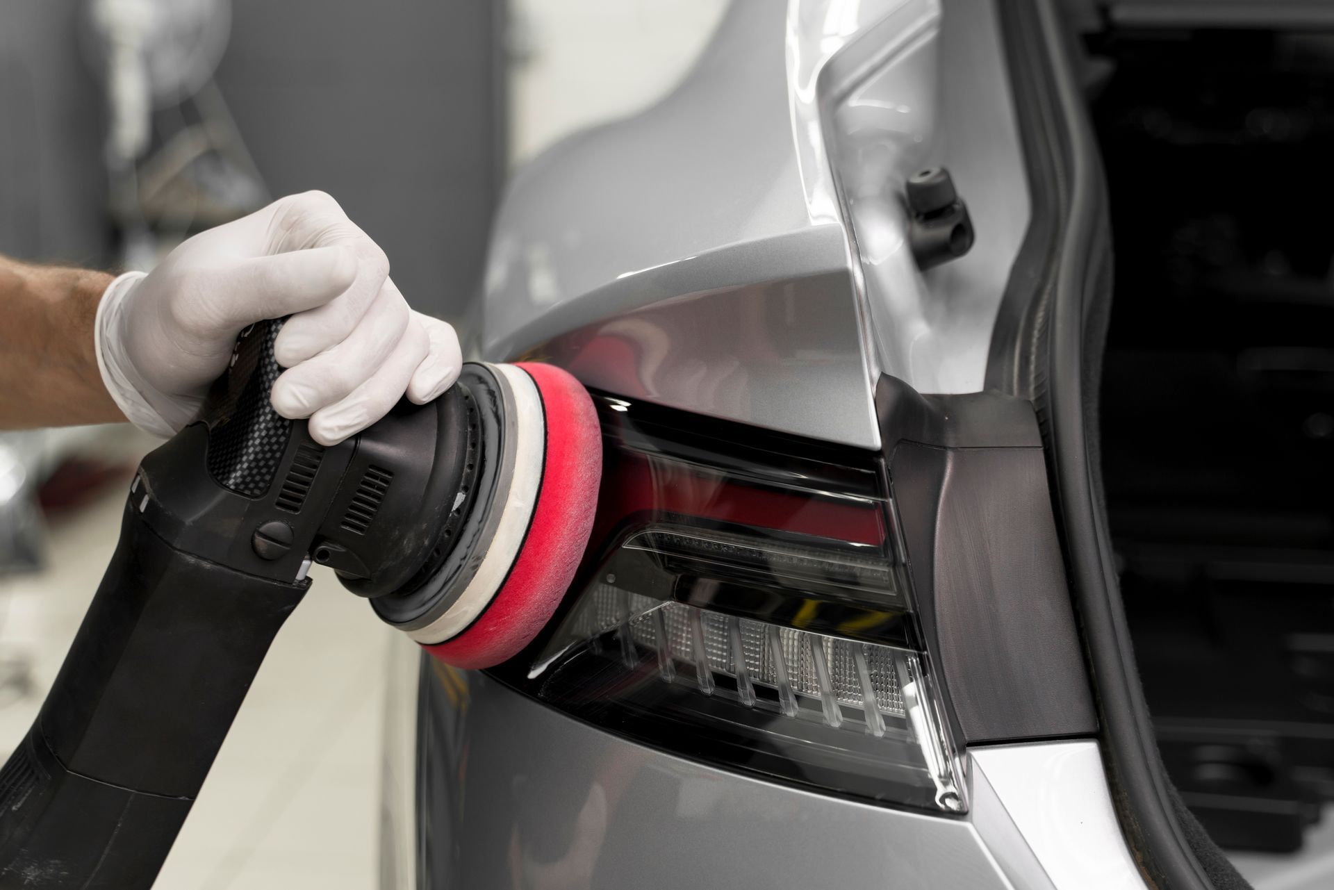 Person using a buffer to polish a car's taillight. The car is silver. The person wears gloves.