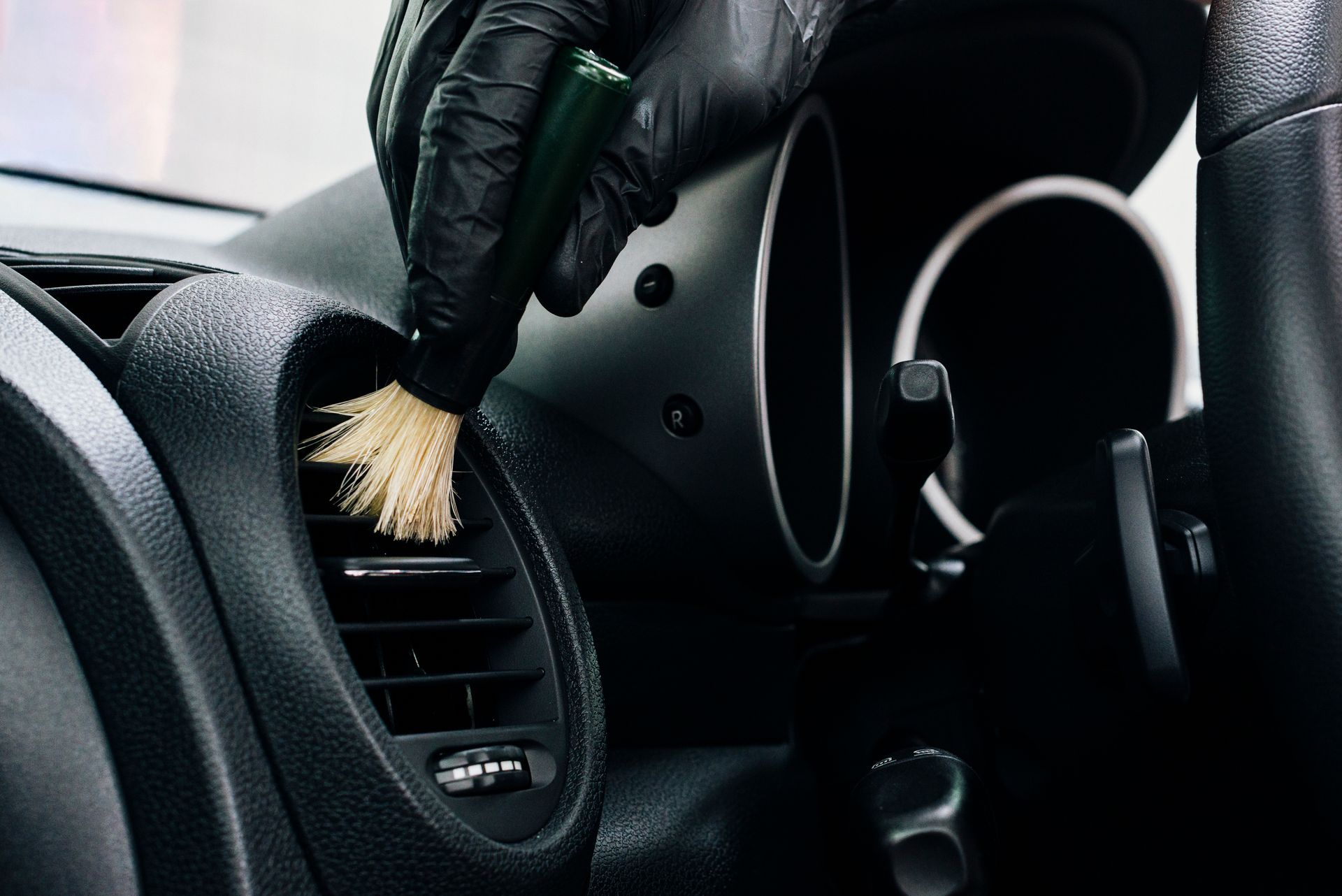 Gloved hand dusting a car air vent with a brush; close-up shot of car interior.